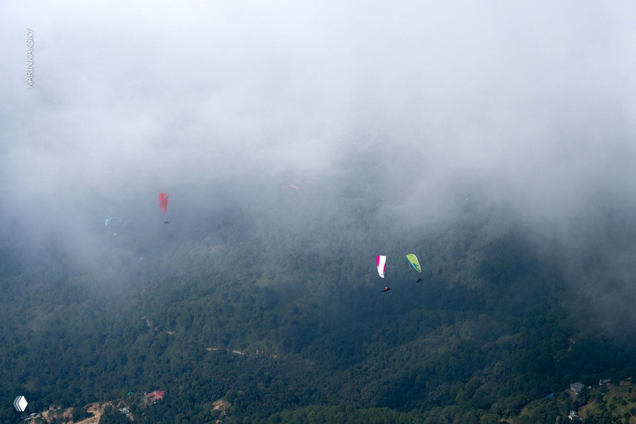 Несколько парапланов плывут над рассеянным туманом и лесистыми склонами; облачный покров снизу и разноцветные крылья пилотов.