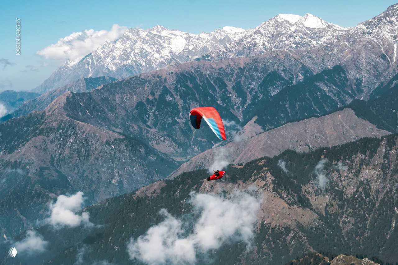 Красный параплан на фоне горных хребтов и склонов; пилот держит высоту над рассыпью облаков и крутыми вершинами.
