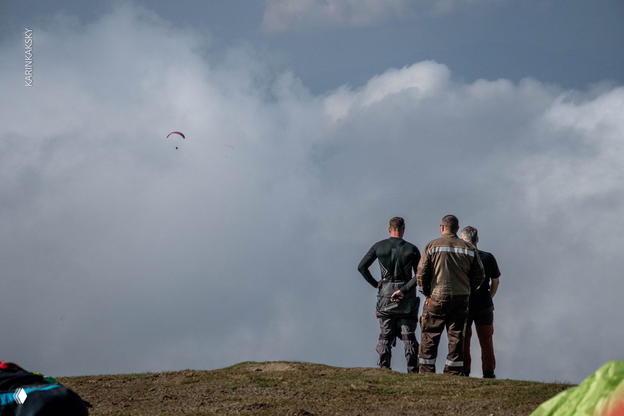 Три человека стоят на краю горного старта и наблюдают за полётом парапланов над морем облаков и долиной внизу.