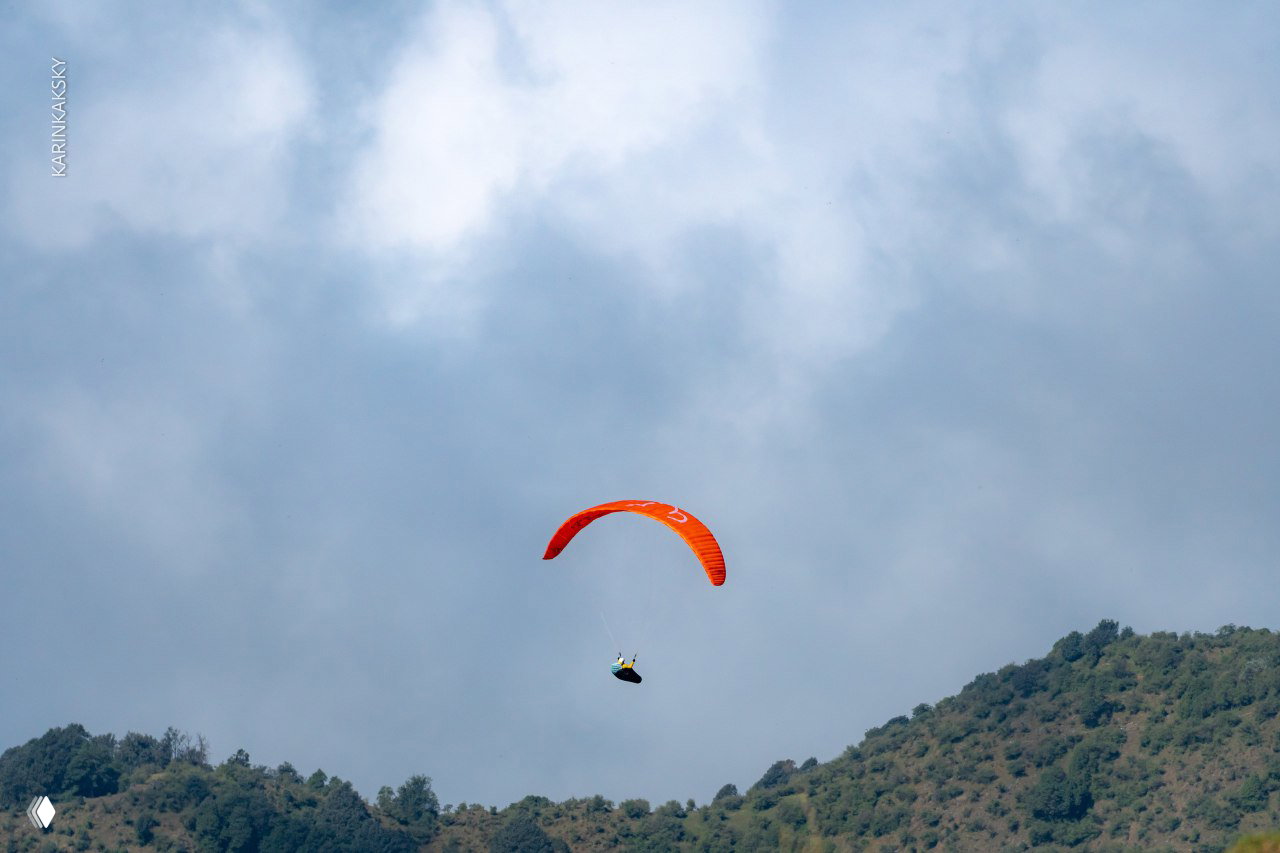 Оранжевый параплан планирует над низким холмом на фоне плотного облачного неба; виден силуэт пилота и зелёные склоны.