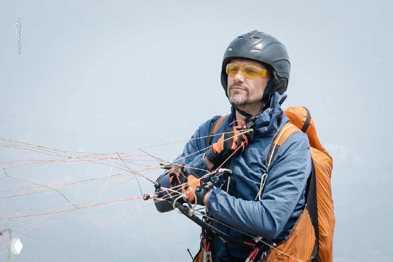 Инструктор в шлеме и желтых очках держит управление парапланом; видно снаряжение и сосредоточенное лицо пилота.