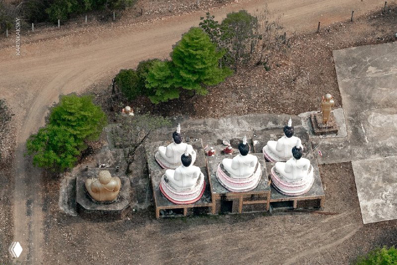 Вид сверху на группу небольших белых статуй Будды на площадке среди сухой почвы и деревьев — ракурс с полёта.
