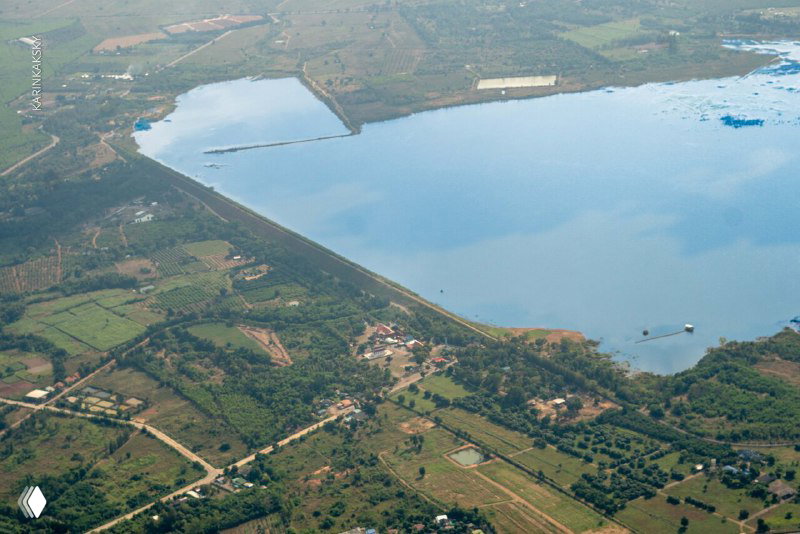 Аэрофотоснимок плотины и водоёма с прилегающими полями и селением; типичный сельский пейзаж Таиланда с высоты.