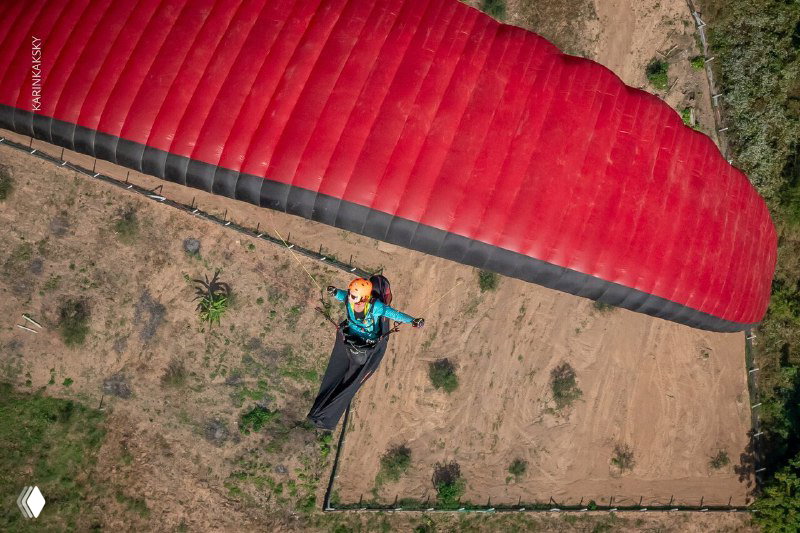 Вид сверху: красное крыло параплана и пилот над покрытой полями землёй, снимок с дрона во время полёта, подчёркивающий масштаб и рельеф местности.