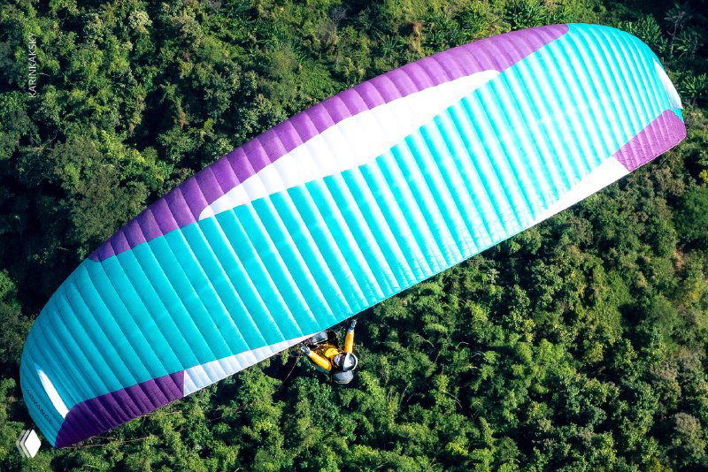 Бирюзово‑фиолетовое крыло параплана над зелёными джунглями; фотография показывает цветное оборудование и густой растительный покров под крылом во время полёта.