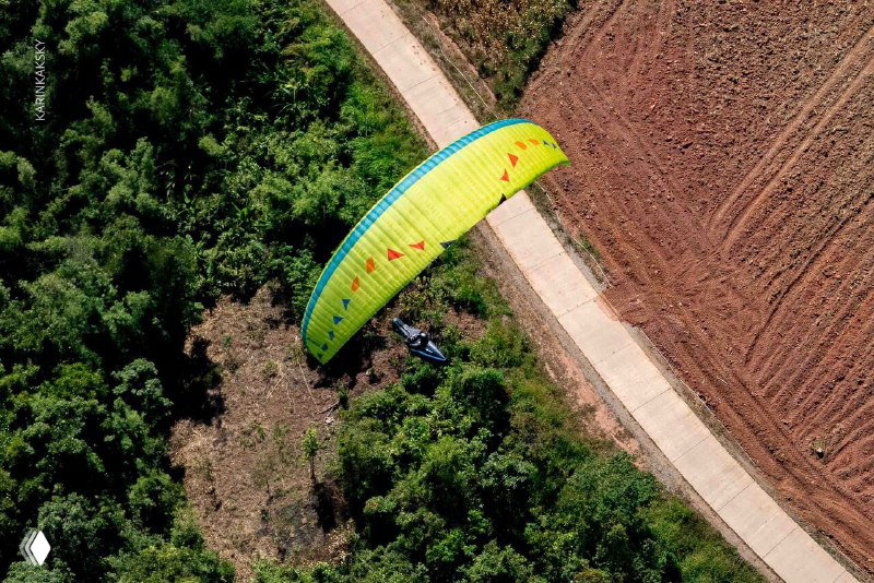 Зелёно‑жёлтый параплан в воздухе над сельским ландшафтом Таиланда, видна дорога и поля снизу — кадр с высоты подчёркивает маршрут и посадочные зоны.