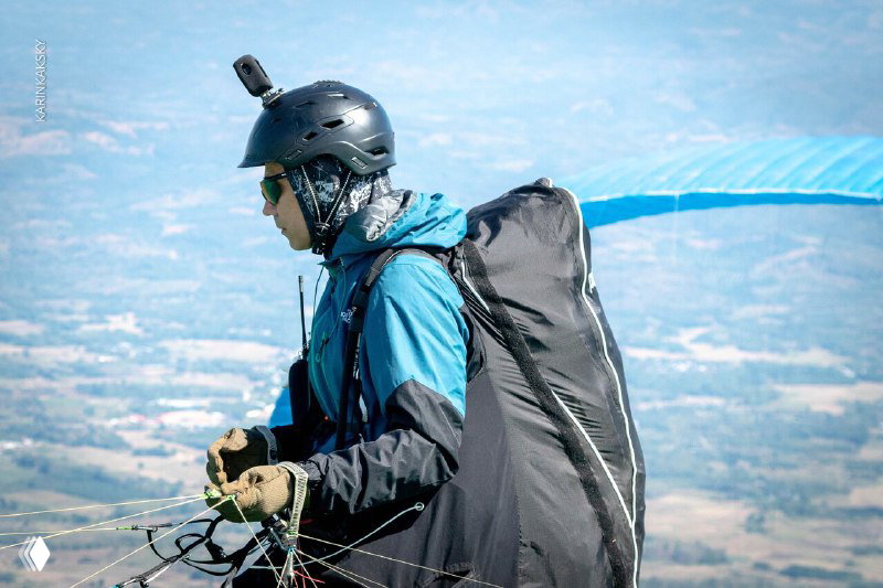 Пилот в подготовке к старту: парапланерист в шлеме и амуниции смотрит вдаль на фон ландшафта и долины перед взлётом.