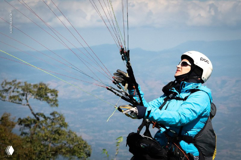 Парапланерист в солнечных очках и шлеме управляет планером на фоне гор и неба, видно управляющие стропы и скалистый пейзаж.