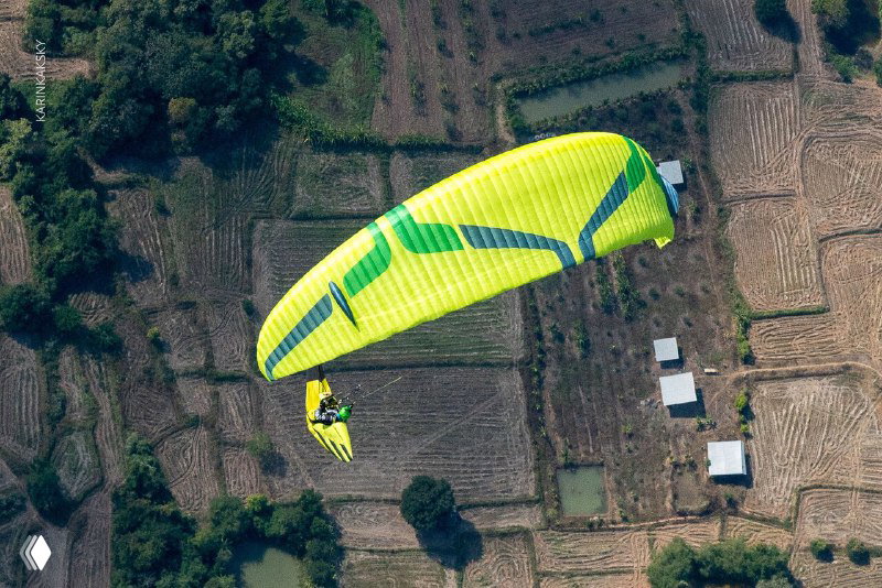 Жёлто-зелёный параплан крупным планом в воздухе, на фоне размытого поля, хорошо видно профиль крыльевого купола и подвесное сиденье.