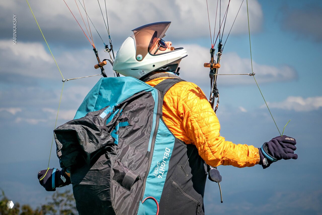 Парапланерист в жёлтой куртке и шлеме, снятый крупным планом во время полёта; видны стропы, рюкзак и детали подвесной системы.