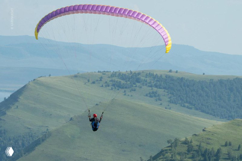 Парапланерист в подвесе над холмами и лесистыми склонами, видна форма крыла и спокойный летный пейзаж для маршрутных полётов.