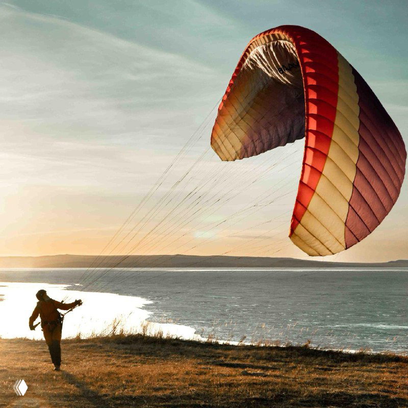 Крупный план купола параплана у водоёма на рассвете; игра света на ткани и подготовка к запуску или приземлению у берега.