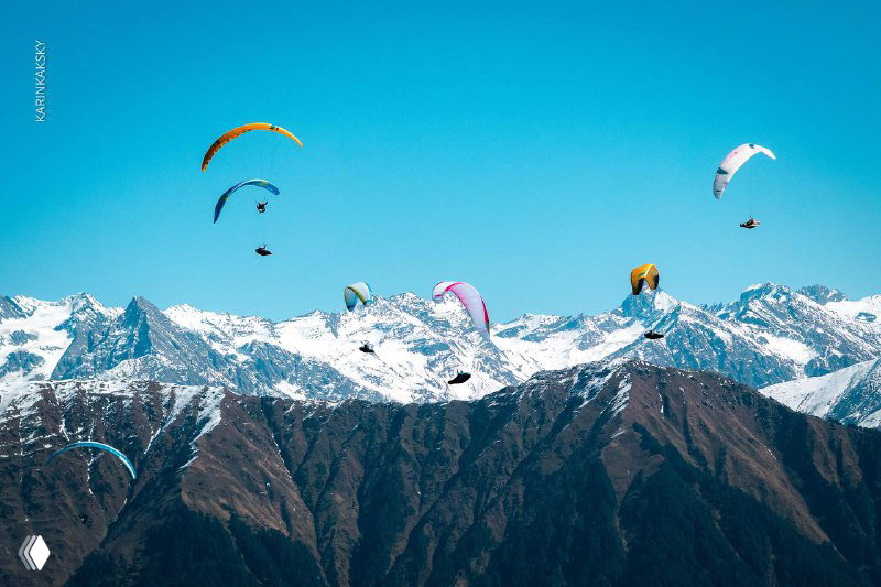 Несколько парапланов в небе над высокими горными пиками и заснеженными хребтами, массовый старт на фоне альпийского ландшафта.