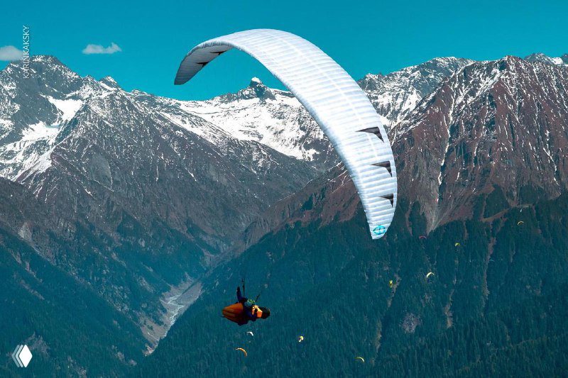 Параплан на фоне высоких заснеженных гор; пилот в подвесе и ясная горная погода подходящая для маршрутных полётов и тренировок.