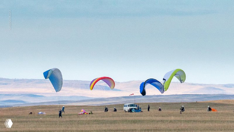 Несколько парапланов на открытом поле Хакасии: разноцветные крылья, люди и машина на заднем плане во время тренировочного дня.