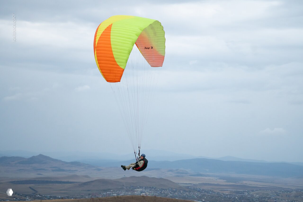 Пилот в подвесной системе на фоне далёких холмов и неба, парящий на ярком зелёно-оранжевом параплане во время полёта.