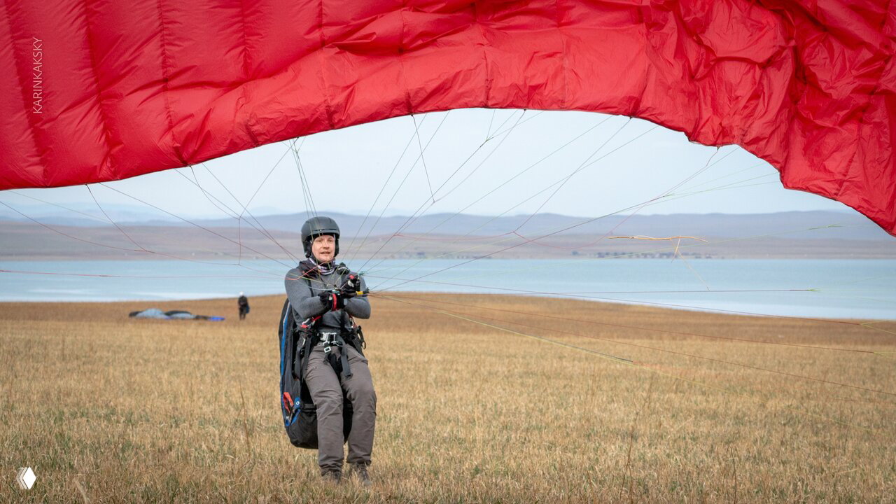 Пилот на ровном поле с поднятым красным парапланом, видна прибрежная зона и спокойная атмосфера тренировочного дня в Хакасии.