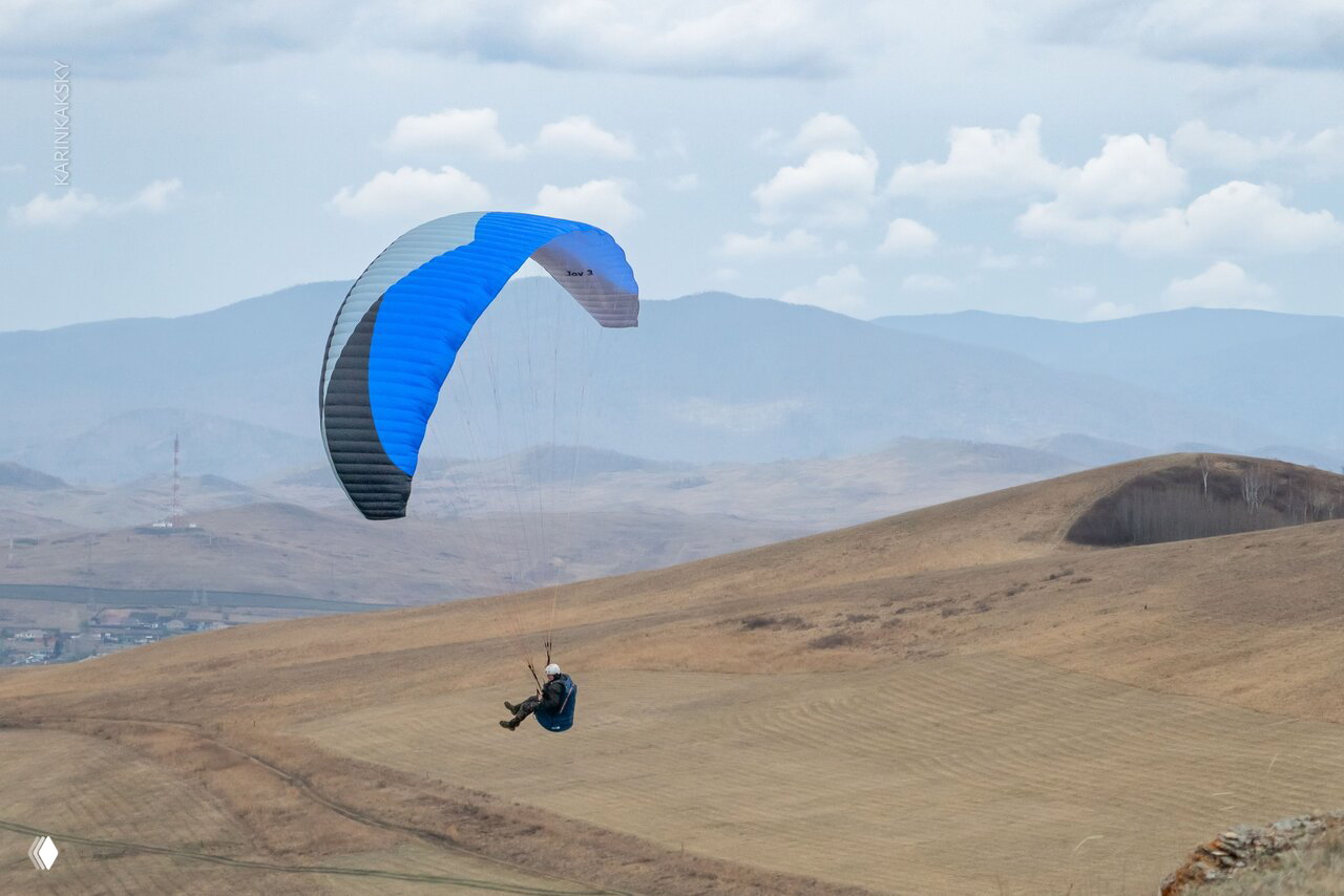Парапланист в воздухе на синём и сером крыле летит над холмами и равниной, демонстрируя обучение термическим и маршрутным полётам.