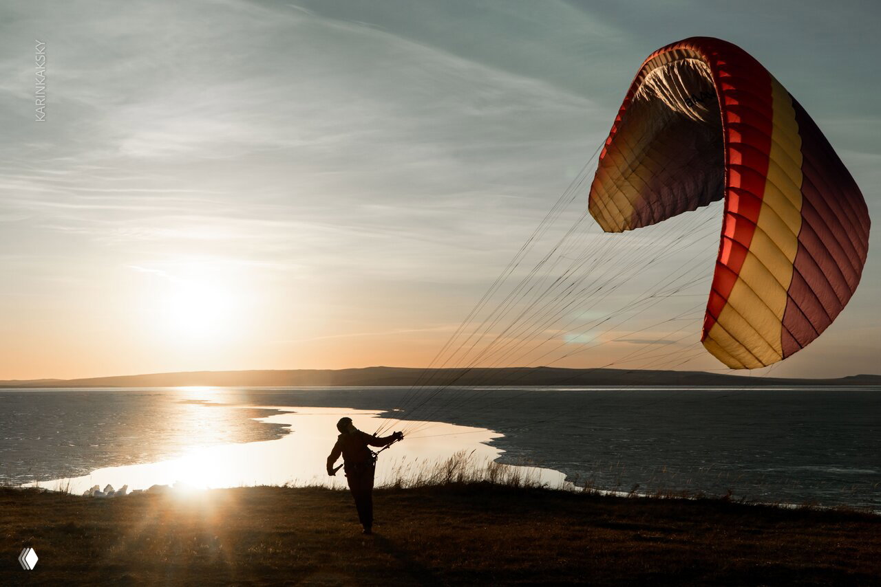 Пилот стоит на утёсе на фоне заката и воды, держит параплан частично над головой — живописный момент тренировочного полёта при закате.