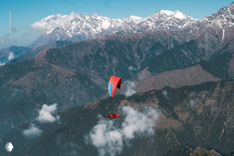 Красный параплан с пилотом над горным хребтом и облаками — панорамный воздушный кадр, демонстрирующий мастерство в полёте.