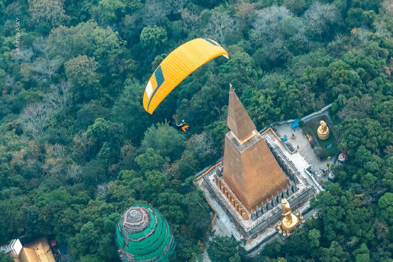 Аэрофотосъёмка жёлтого параплана над густым лесом и крупной пагодой с конической ступой, вид сверху, Тайланд.