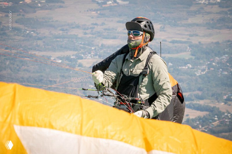Пилот в шлеме и солнцезащитных очках держит управление парапланом, видна передняя кромка жёлтого купола в ближайшем плане.