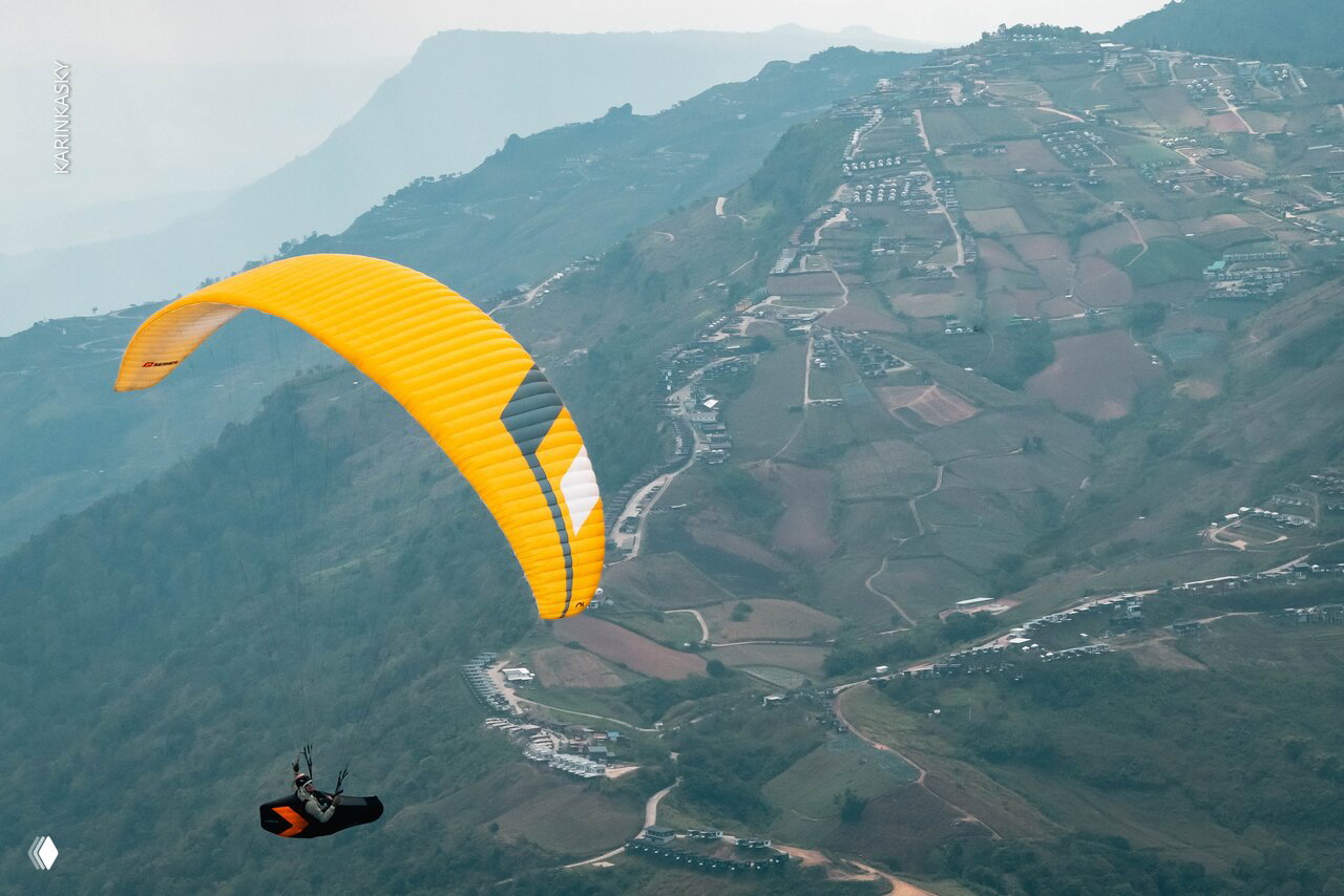 Жёлтый параплан в широкой горной долине: вид с высоты показывает поселения, дороги и террасы в глубине пейзажа.