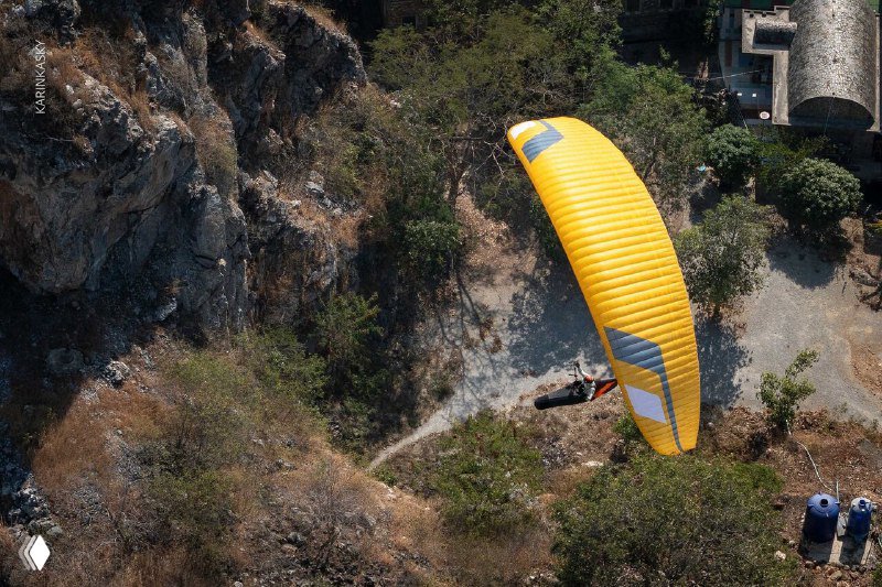 Параплан снижается над скалистой местностью и редкой растительностью, пилот виден под куполом над посадочной зоной.