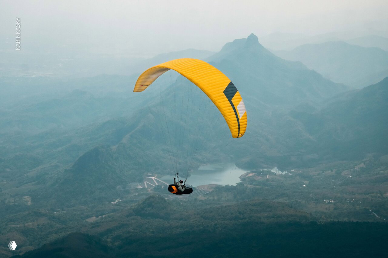Жёлтый параплан в полёте над холмами, вдалеке видна коническая гора и небольшое озеро — атмосферный пейзаж Таиланда.