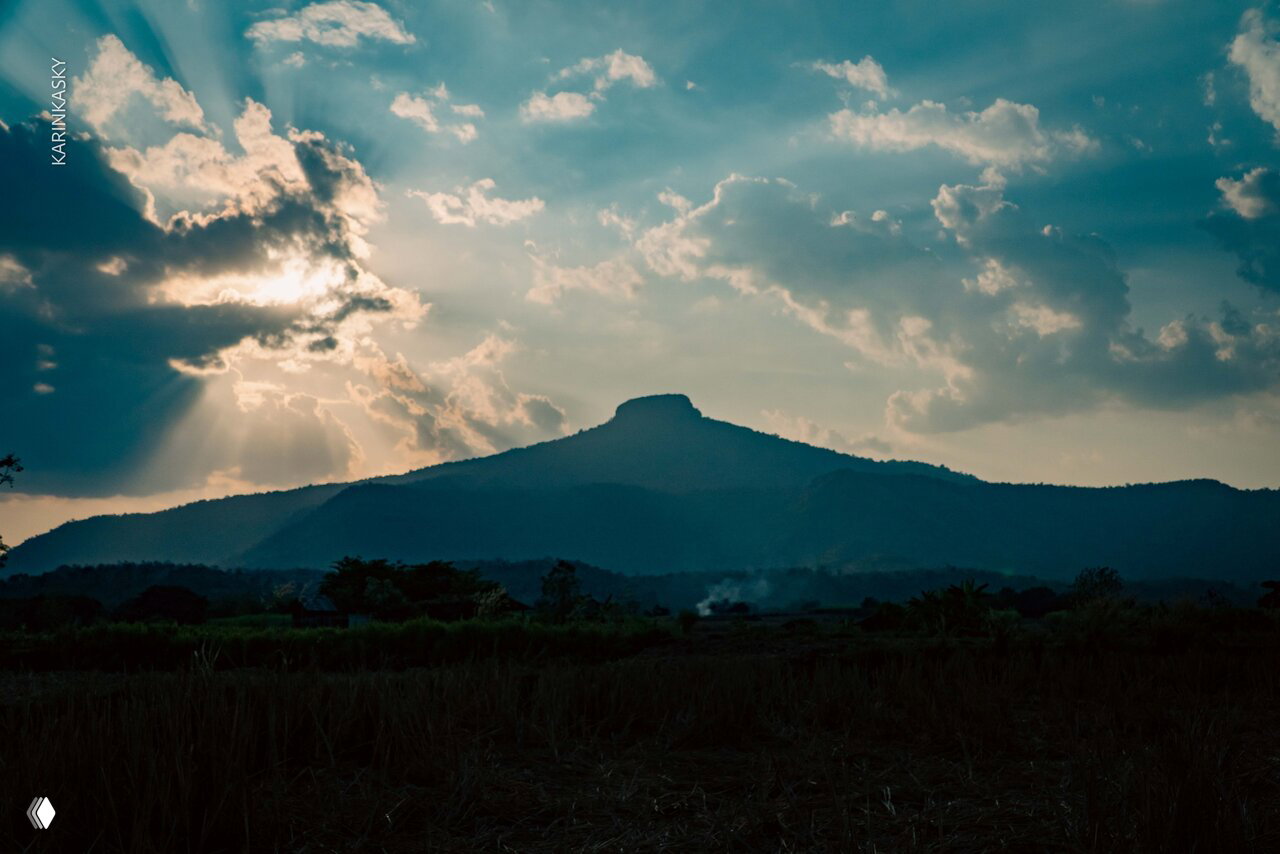 Драматичный пейзаж с конической горой на фоне неба и лучей солнца, передний план — тёмная равнина и лёгкий дымок.