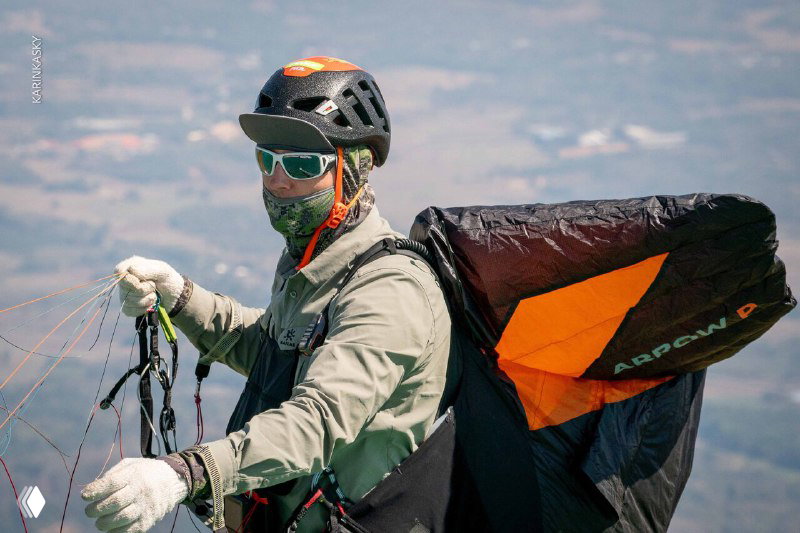 Пилот в маске и шлеме держит стропы параплана в полёте, хорошо видны снаряжение, подвесная система и элементы управления.