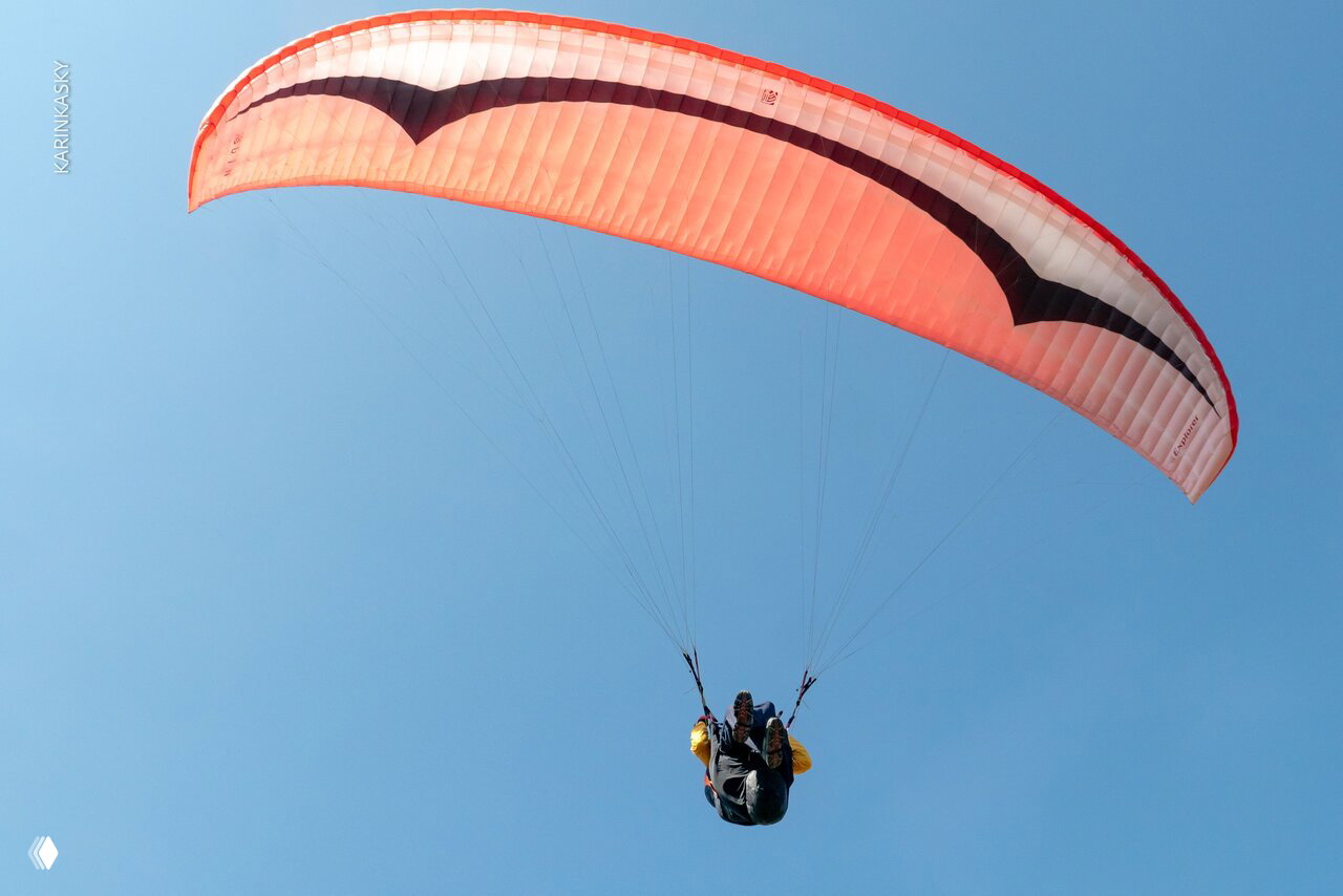 Светло‑оранжевое крыло параплана на фоне чистого голубого неба в Таиланде; низкий ракурс показывает форму купола и подвеску пилота в полёте.