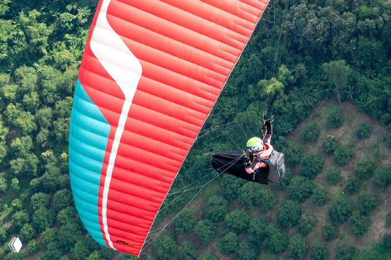 Параплан в полёте над густой тропической растительностью; вид сверху на красно‑бирюзовое крыло и спускающиеся джунгли внизу, обширный ландшафт.