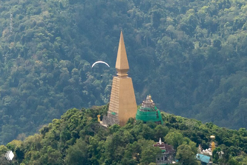 Золотая пагода на вершине зелёного хребта; в небе позади виднеется параплан — сочетание местной архитектуры и полётов во время выезда в Таиланд.
