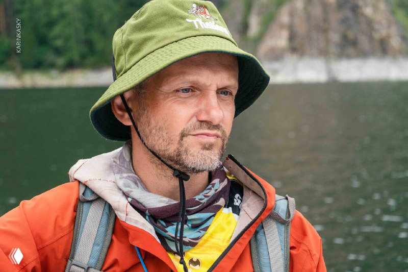 Портрет пилота AirSiberia в походном снаряжении на фоне воды и лесов, спокойное лицо и защитный головной убор для полётов.