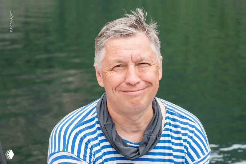 Портрет ещё одного члена команды AirSiberia у воды: улыбающееся лицо, ветровка и фон с лесистой береговой линией и горами.