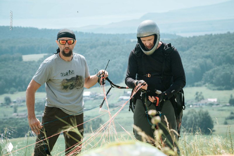 Два пилота готовят параплан к запуску на склоне: контроль строп, шлемы на участниках и вид сельской долины внизу.