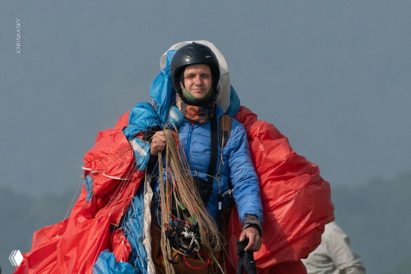 Пилот идёт с ярким сложенным парашютом на спине: экипировка, рюкзак и следы подготовки после приземления на фоне равнин.