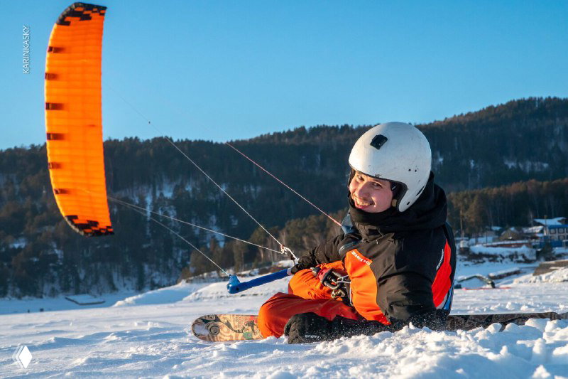 Кайт-скейтинг зимой: человек отдыхает на снегу с ярким кайт-крылом, зимний пейзаж и горы на заднем плане.