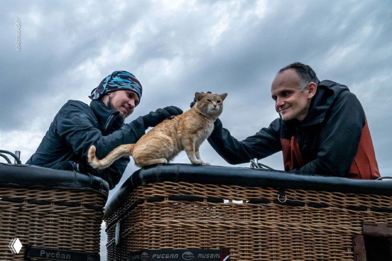 Корзина воздушного шара с двумя пилотами и котом, уютная сцена посадки или подготовки аэростата на фоне облачного неба.