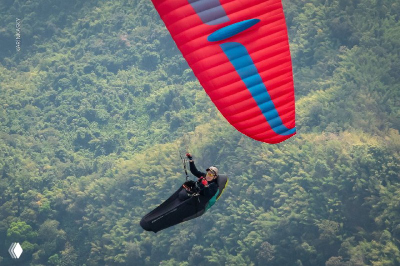 Близкий план: пилот на красном параплане в подвесной системе на фоне зелёного горного склона — момент уверенного полёта и работы в потоках.