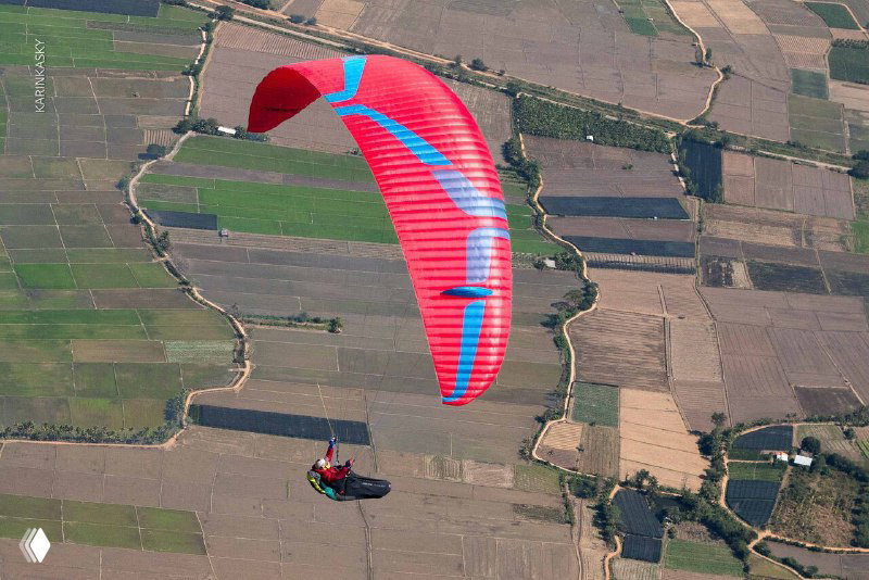Красный параплан в полёте над мозаикой полей и сельхозучастков — воздушная перспектива маршрутов и живописный ландшафт.