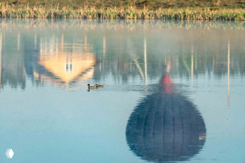 Отражение воздушного шара и домиков в спокойной глади озера, на переднем плане пара уток; мягкий рассветный свет и зеркальная вода.