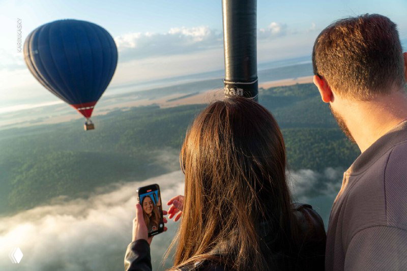 Вид из корзины воздушного шара: женщина и мужчина делают снимок на телефон, вдалеке другой шар над лесом и утренние облака.