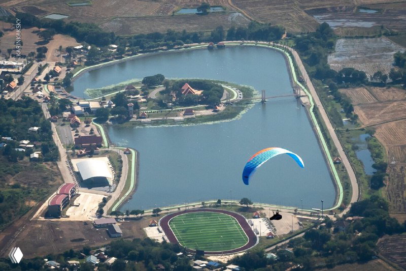 Парапланист над водоёмом и парком Таиланда: вид сверху на ландшафт, водную гладь и одиночный цветной параплан в воздухе.