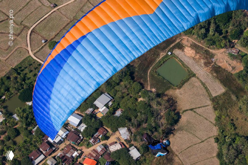 Синий и оранжевый купол параплана крупным планом над сельской частью Таиланда, видны дома и сельхозучастки под крылом.