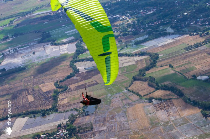 Зелёный парапланист летит над холмами и полями Таиланда: подвесной пилот виден на фоне разреженной застройки и сельхозугодий.