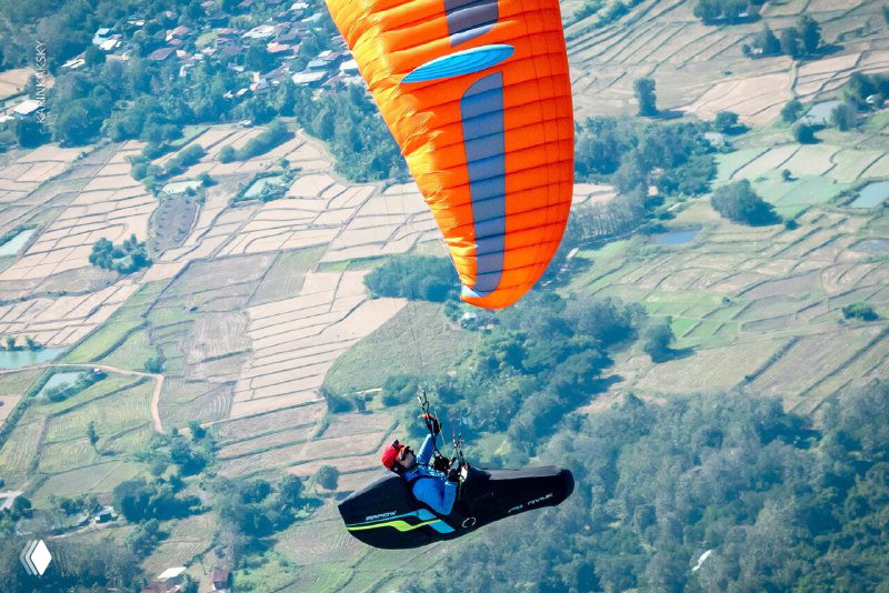 Оранжево-синий параплан над террасными полями и холмами Таиланда, пилот в подвесной системе виден на фоне пейзажа.