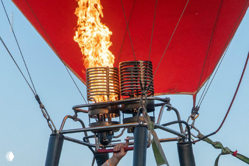 Крупный план газовой горелки воздушного шара с открытым пламенем: металлическая рама, трубопроводы и факел пламени, снимаемая снизу.