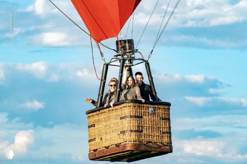 Пассажиры в плетёной корзине воздушного шара на фоне голубого неба: люди стоят в люльке, видны канаты и низ оболочки шара.
